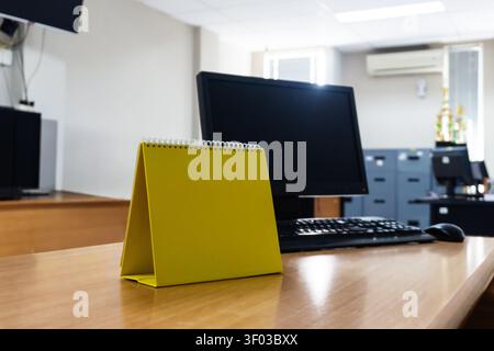 Bright yellow calendar sits on wooden desk. Black monitor and keyboard nearby, office environment. Soft lighting, tidy setup suggests professionalism. Ideal for business, planning Stock Photo