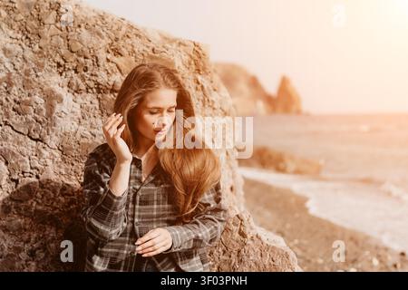 Woman beach rocks young female standing by stone cliff at seashore looking down thoughtfully in soft evening sunlight Stock Photo