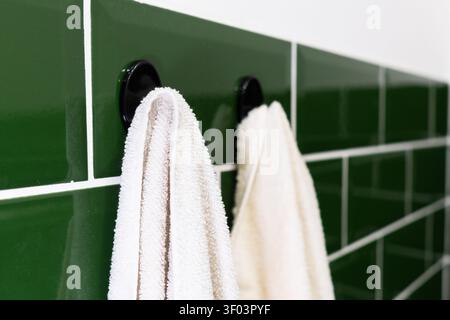 White cotton towels hanging on black plastic hooks on dark green ceramic tile wall background in bathroom. High quality Stock Photo