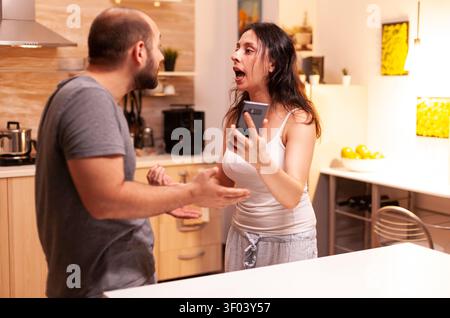 Angry wife confronts husband over cheating messages on a smartphone, showing infidelity, jealousy, frustration and relationship conflict Stock Photo