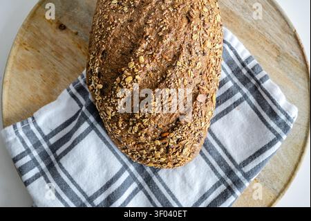 Overhead view of a seeded whole grain bread loaf resting on a checkered cloth and wooden board, a rustic artisan loaf covered with oats and seeds, a w Stock Photo