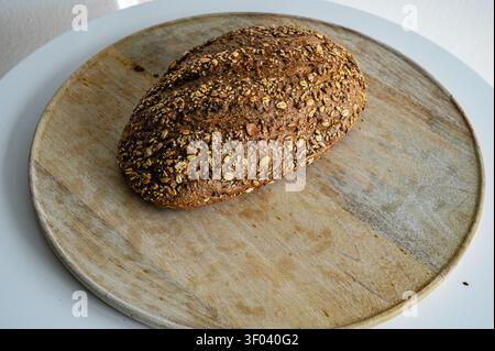 Artisan multigrain bread loaf with a golden seeded crust resting on a round wooden board, a wholesome homemade loaf full of grains against a soft back Stock Photo