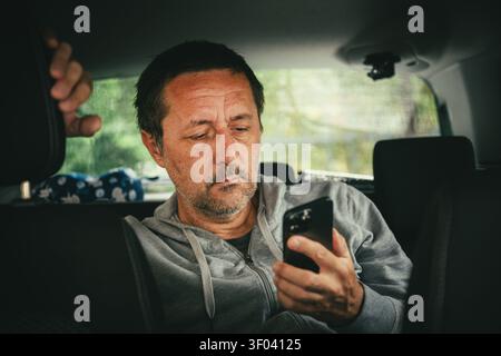 Middle-aged man looking at smartphone while sitting in car with concerned expression. Selective focus Stock Photo