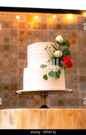 A two-tier white wedding cake with textured buttercream is topped with white and burgundy peonies flowers on a silver stand with string lights behind Stock Photo