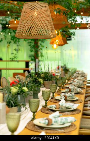 A long outdoor dining table is styled with green glassware, floral-patterned plates, greenery runner and a woven pendant lamp under string lights. Stock Photo