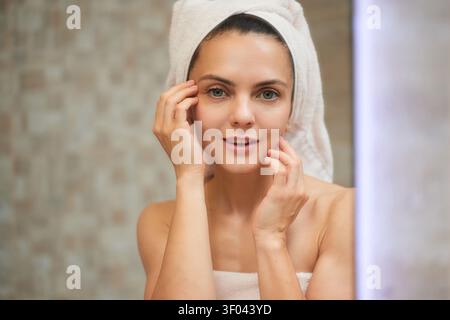 Woman standing in bathroom with towel on head and wrapped around body, touching her face, applying facial cream Stock Photo