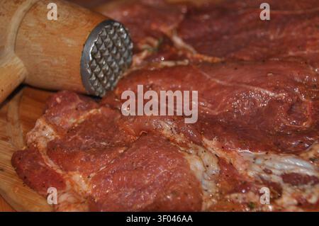 Fresh, marinated meat is placed on a wooden board. A meat tenderizer is used to pound the meat, preparing it for cooking. The scene showcases kitchen Stock Photo