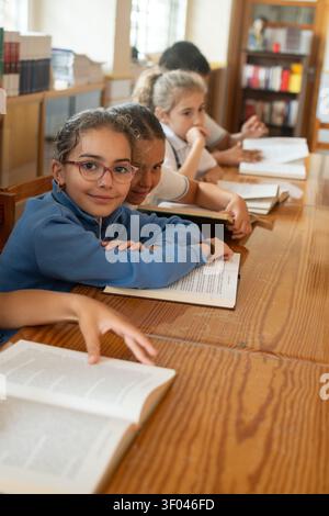Smiling Elementary School Girl Reading with Classmates in a School Library Stock Photo