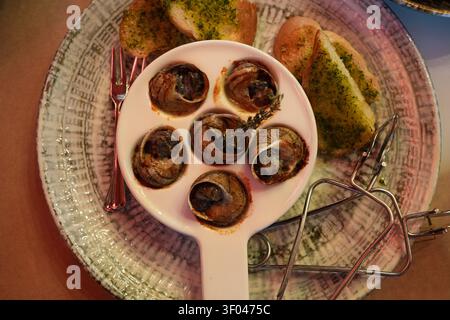 Top view of baked escargot snails in a white ceramic dish with herb toast and serving tongs. Fine French cuisine and elegant restaurant plating. Stock Photo