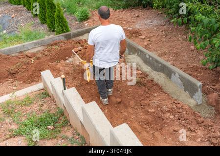 View from behind of a man pushing a wheelbarrow on dirt ground at a residential construction site. Building a modern concrete block retaining wall for Stock Photo