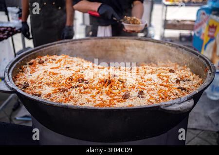 Central Asian pilaf being served from a large black cauldron at an outdoor street food market. Traditional rice dish, festive catering, and hot meal. Stock Photo
