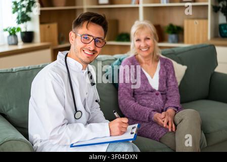 A friendly healthcare professional smiles while taking notes during a home visit to an elderly female patient. Stock Photo