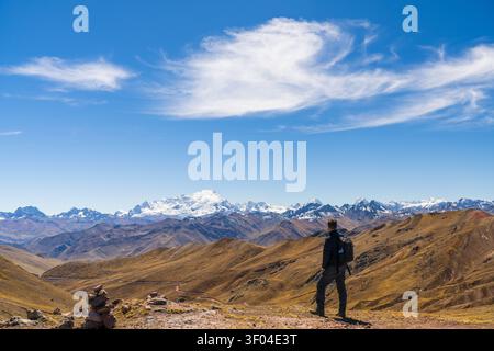 A hiker stands on a ridge near Palccoyo Rainbow Mountain, looking at the snow-capped peak of Mount Ausangate under a bright blue sky, Peru Stock Photo