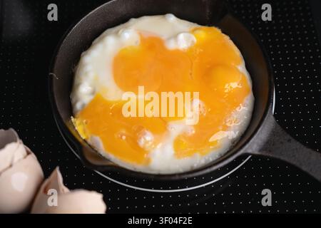 overhead close-up of cracked eggs frying in a small castiron skillet on a black dotted induction cooktop, glossy vibrant yellow-orange yolks pooling o Stock Photo