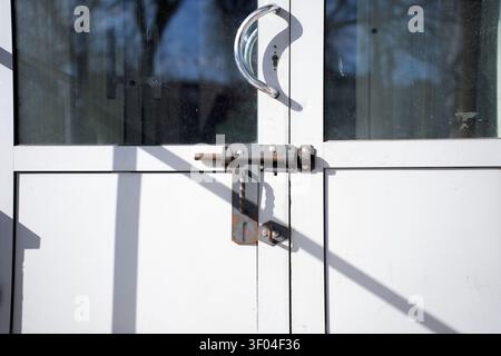 close-up of a white metal double-door entrance in daylight featuring a chrome curved handle, rusted sliding-bolt latch, dusty glass panes and strong d Stock Photo