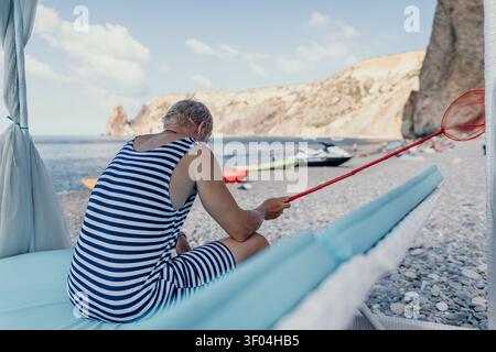 Beach man net mature man wearing a striped swimsuit sits on a lounge chair holding a red butterfly net on a rocky coastline Stock Photo
