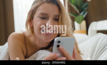 Satisfied cute pretty Caucasian woman waking up in bed receive mobile phone message chatting with online app sms smiling lying in bedroom at morning Stock Photo