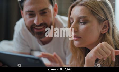 Optimistic Caucasian family couple positive talking using digital tablet network using internet laughing lying in bedroom in morning at home European Stock Photo