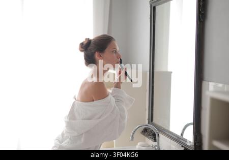 Young woman in bathrobe applying makeup in mirror Stock Photo