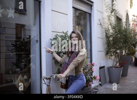 Smiling woman on a bicycle pointing at a store Stock Photo