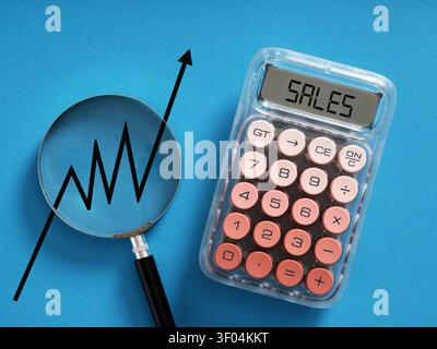 Sales word displaying on calculator screen next to magnifying glass showing an upward sales trend line on a blue background. Increasing sales concept. Stock Photo