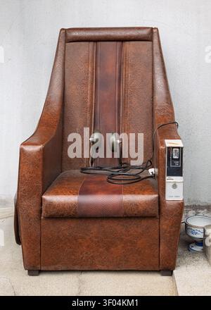 Vintage brown leather coin-operated massage chair standing indoors against a plain background wall, Kyoto, Japan Stock Photo