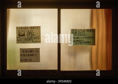 Dark night view of illuminated window panes displaying white paper notices with printed Japanese text, Kyoto, Japan Stock Photo