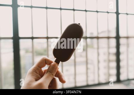 Hand Holding Chocolate Ice Cream Bar in Front of Large Industrial-Style Window in Berlin, Germany Stock Photo