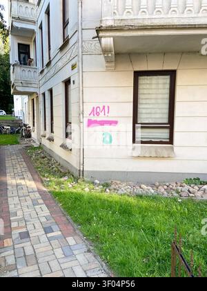 Flensburg, Schleswig-Holstein Nahaufnahme einer hellen Putzfassade eines Mehrfamilienhauses an einer Straßenecke in Flensburg. Auf der Wand ist ein auffälliges, mit rosa Sprühfarbe aufgetragenes Graffiti zu sehen, das den Zahlencode 161 symbolisch für AFA - Antifaschistische Aktion zeigt. Darunter befindet sich ein übermalter Bereich und ein kleines, grünes Logo-Symbol. Die Szenerie zeigt einen gepflasterten Gehweg, einen Rasenstreifen mit Steinen und einen Blick in den Hinterhof mit abgestellten Fahrrädern. Ein Fenster mit heruntergezogenen Jalousien befindet sich rechts vom Graffiti. Der Zus Stock Photo