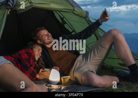 Two hikers relax in their tent at dusk after a day of trekking in the mountains. They smile while taking a selfie together. Stock Photo