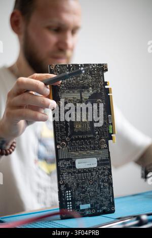 Man works on a computer graphics card while holding it in one hand and using tools with the other hand in a well-lit room Stock Photo