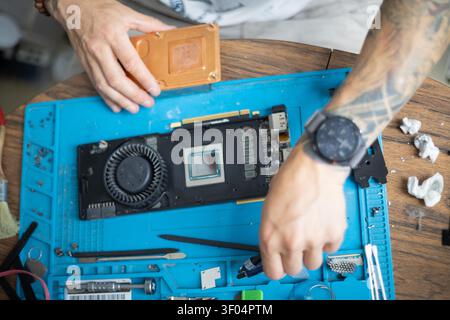 Person repairs a graphics card on a blue work surface with various tools and parts around in a home setting during daytime Stock Photo