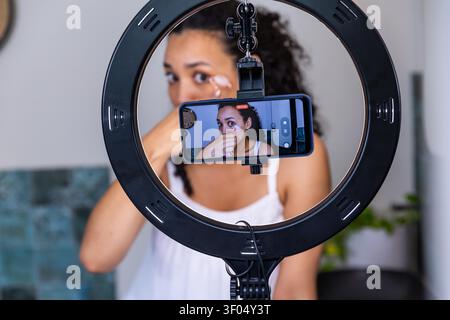 Adult African American woman in tank applying undereye cream filming on ring light phone at vanity. Skincare, makeup, beauty, demonstration, creator, circular, led Stock Photo