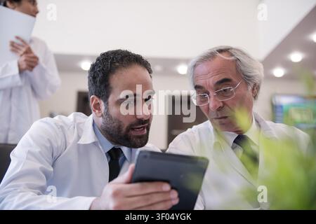 Two doctors in lab coats review patient data on a tablet while discussing an upcoming eye surgery. The scene conveys focused teamwork, mentoring, and professional ophthalmology care in a small clinic setting. Stock Photo