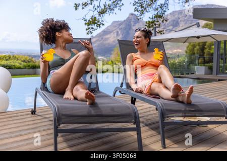 Diverse female friends reclining on pool deck lounge chairs in bikinis, holding duck cups, talking. Women, outdoor, sunlit, terrace, modern, relaxation, mountains Stock Photo