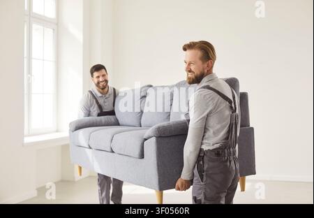 Two young moving service employees workers carrying heavy sofa together in the living room Stock Photo