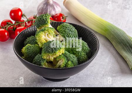 Vibrant green broccoli florets in textured black bowl surrounded by red cherry tomatoes garlic bulb leek on light gray surface. Natural lighting highl Stock Photo