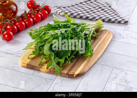 Fresh green arugula on wooden board beside red cherry tomatoes olive oil bottle. Gray marble countertop with checkered napkin. Natural light highlight Stock Photo