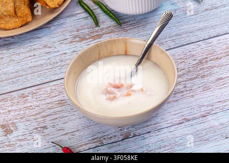 Creamy yogurt in ceramic bowl with scattered nuts spoon, set on weathered blue wooden surface. Green red chilies add color contrast. Soft natural ligh Stock Photo