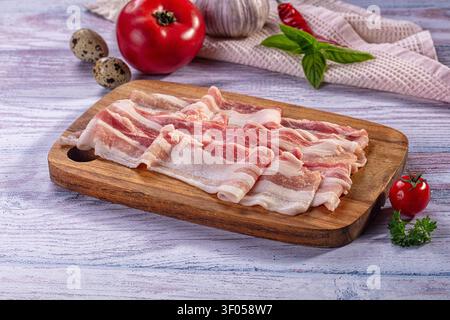 Fresh raw bacon slices arranged on rustic wooden board. Composed with red tomato garlic chili basil quail eggs. Soft natural lighting highlights marbl Stock Photo