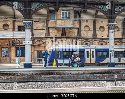 A couple sit on a railway station platform bench and a man stands nearby using a phone. A train is in a siding behind them and a brick wall is in the Stock Photo