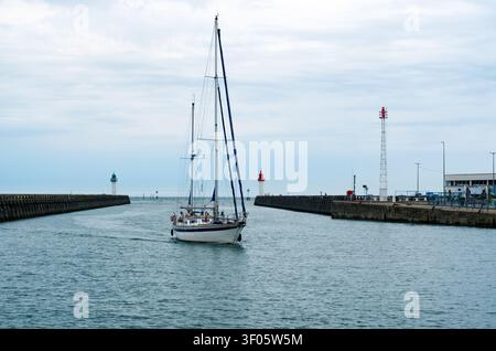A sailboat navigating through a harbor channel toward the open sea, passing between red and green navigation lighthouses on concrete breakwaters. The Stock Photo