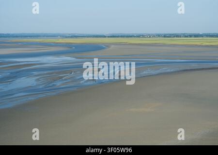 A wide coastal tidal flat with shallow blue water channels winding across smooth wet sand during low tide. Natural patterns formed by water and sedime Stock Photo