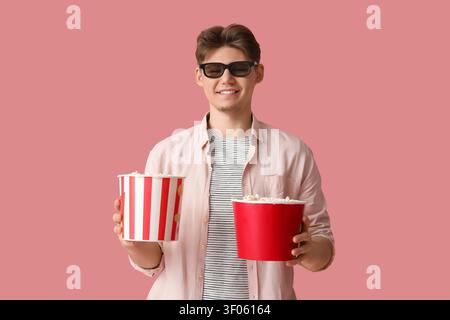 Handsome young man in 3D glasses holding buckets with popcorn on pink background Stock Photo