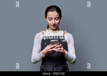 Young female in apron looking down at smartphone screen on gray background Stock Photo