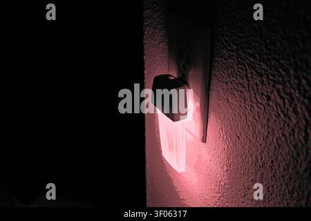 Red night light plugged into a wall outlet glowing in a dark room, suggesting low-light safety, comfort, and ambient home lighting. Stock Photo