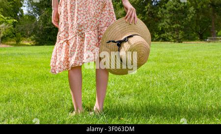A close-up of a young woman standing on a lush green lawn, holding a straw hat in one hand Stock Photo
