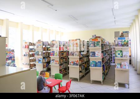 A child sits reading at a colorful table in a bright, spacious library filled with tall bookshelves and children's books Stock Photo