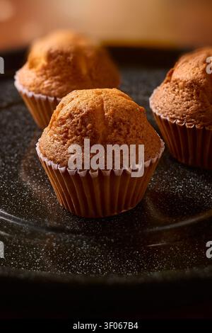 freshly baked golden muffins on dark speckled tray close-up, warm sweet pastry in paper cups, side view with soft focus and copy space, homemade Stock Photo
