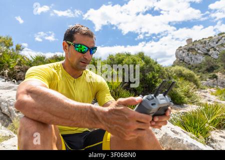 Athletic man in yellow sportswear and mirrored sunglasses operates a drone controller while sitting on rocks, with green shrubs, cliffs, and a bright Stock Photo